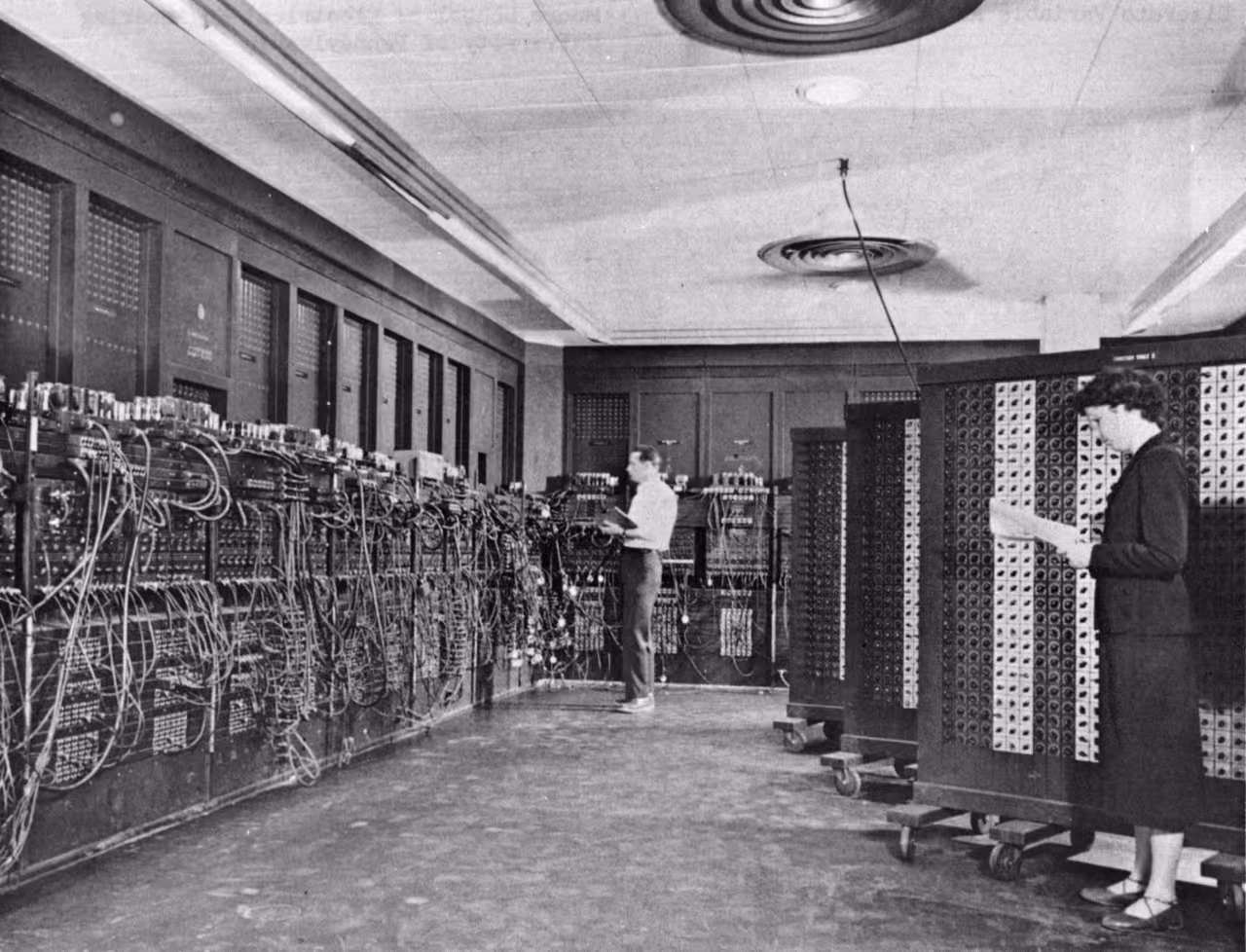 Een historische zwart-witfoto van een kamerbrede vroege computer met talloze kabels en schakelpanelen waar technici aan het werk zijn.