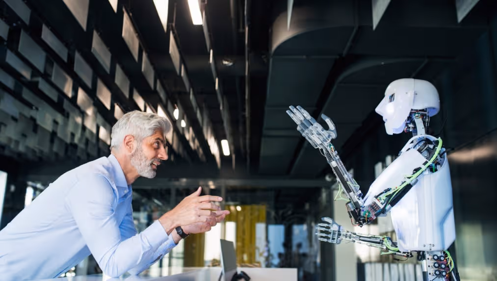 Een grijsharige man in een lichtblauw overhemd die op een balie leunt en een gesprek voert met een witte humanoïde robot in een modern, industrieel kantoor.