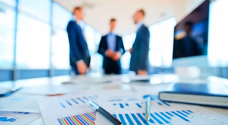 Een wazige achtergrond van drie professionals in pak in een modern kantoor, met op de voorgrond een scherpe focus op grafieken, pennen en een notebook op een witte vergadertafel.
